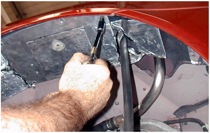 A hand using a tool to remove bolts from a metal pump plate located inside the wheel arch liner of a red car.