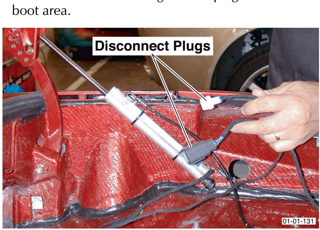 A hand disconnecting two wiring harness plugs located in the red painted boot area of the vehicle.