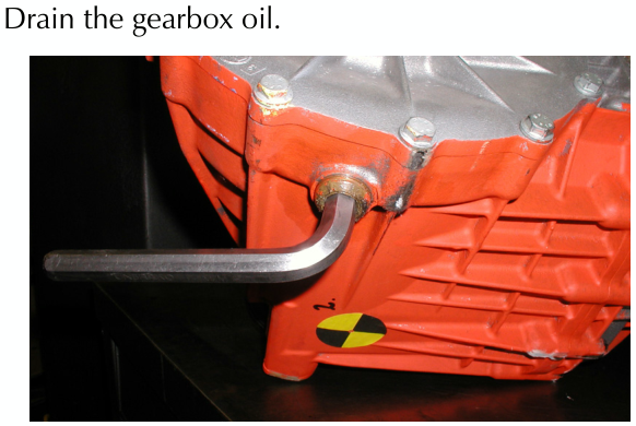 A long L-shaped metal tool inserted into a drain plug hole on an orange differential casing