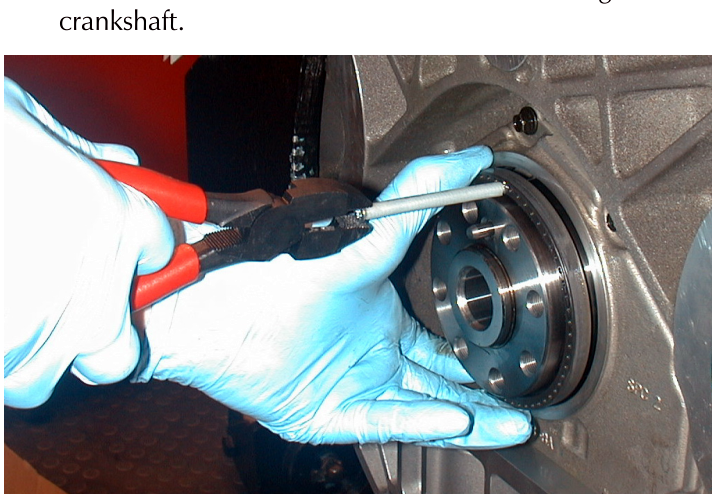 A gloved hand using pliers to pull a circular oil seal off a crankshaft.