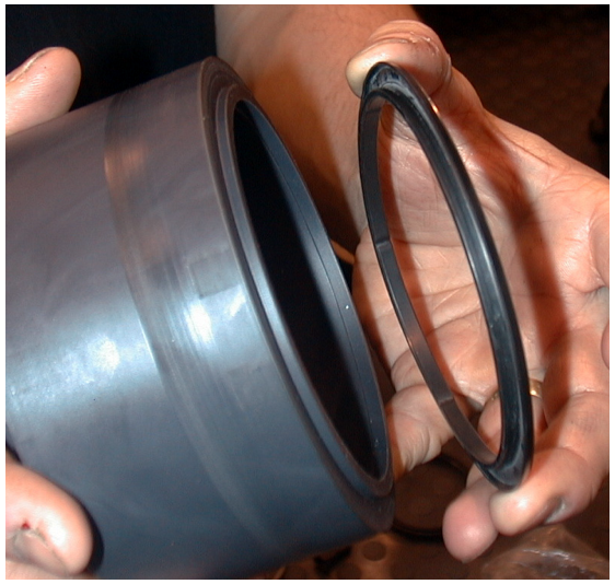 A hand holding a large black cylindrical oil seal slinger next to a thin black rubber O-ring or seal ring.